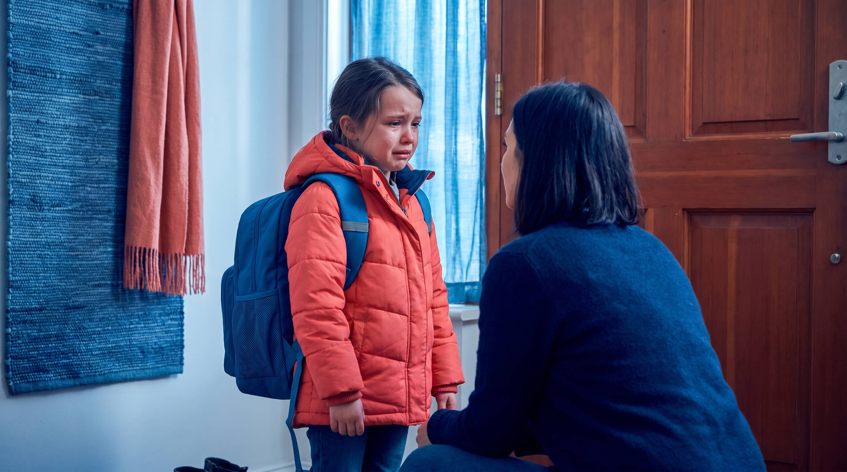 Trauriges Kind mit Schulrucksack mit Mutter in Hausflur